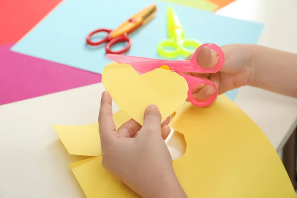 Child cutting out paper heart with plastic scissors at table, closeup