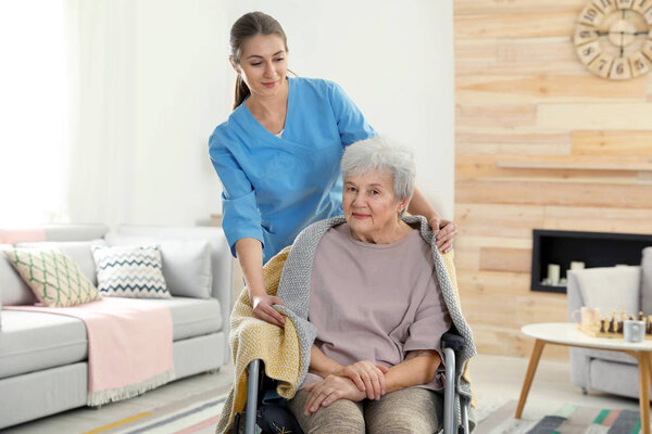 Nurse covering elderly woman in wheelchair with blanket indoors. Assisting senior people