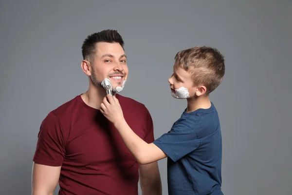 Syn stosując piankę do golenia na twarzy taty, szare tło — Zdjęcie stockowe