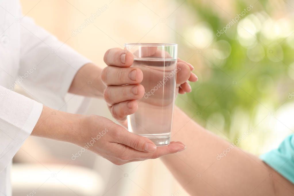 Nurse giving glass of water to elderly man indoors, closeup. Assisting senior generation