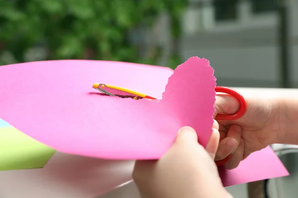 Child cutting out paper heart with craft scissors at table indoors, closeup