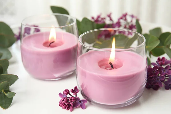 Burning candles in glass holders and flowers with leaves on white table — Stock Photo, Image