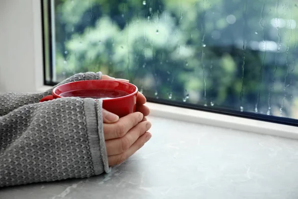 Mujer con taza de té caliente cerca de la ventana en el día lluvioso, primer plano. Espacio para texto — Foto de Stock