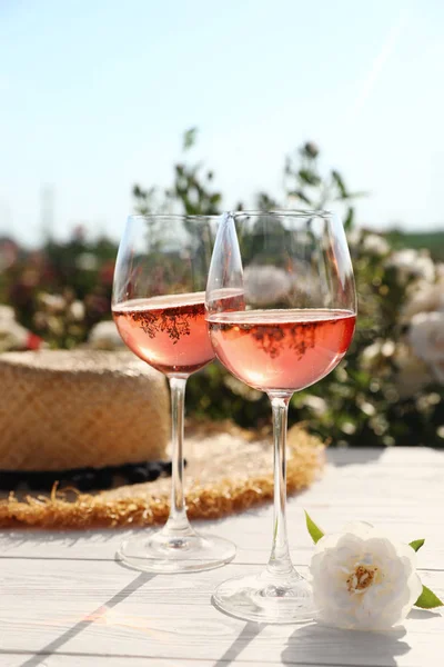 Sklenice růžího vína, slamného klobouku a nádherné květiny na bílém dřevěném stole venku — Stock fotografie