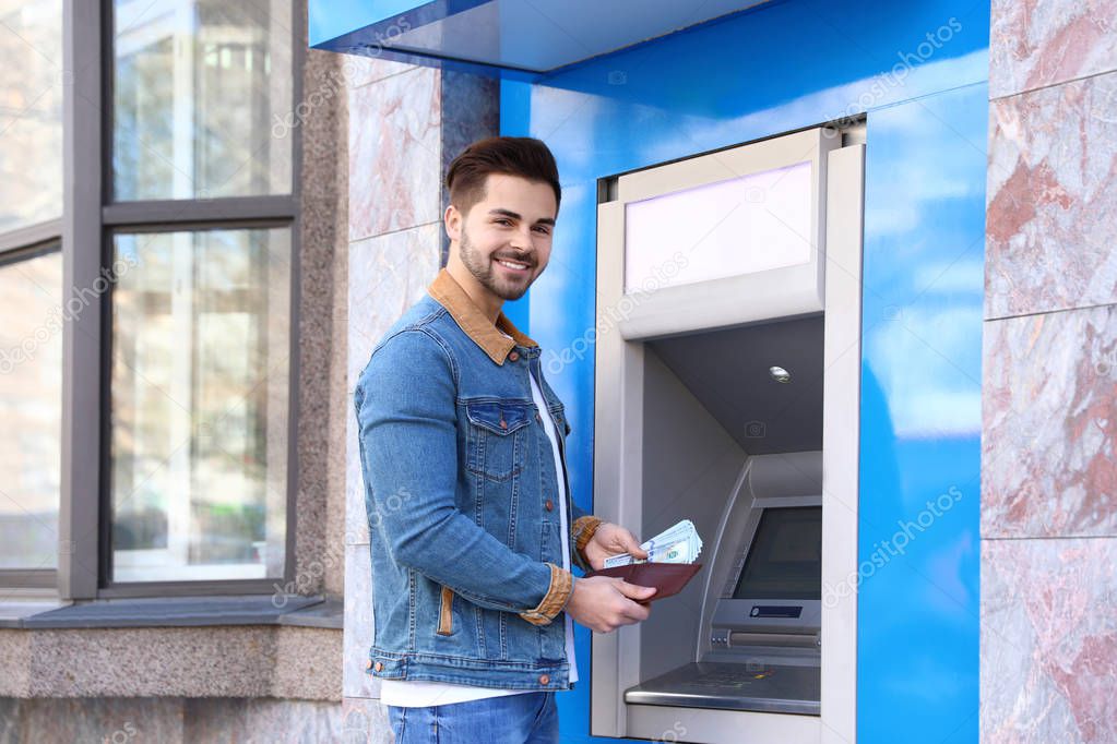 Young man with money near cash machine outdoors