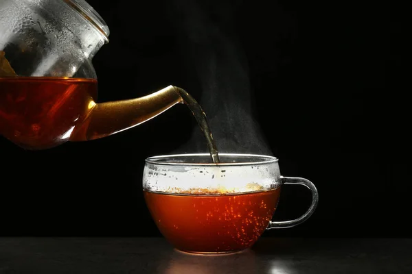 Verter un delicioso té caliente en una taza de vidrio sobre la mesa sobre un fondo oscuro — Foto de Stock