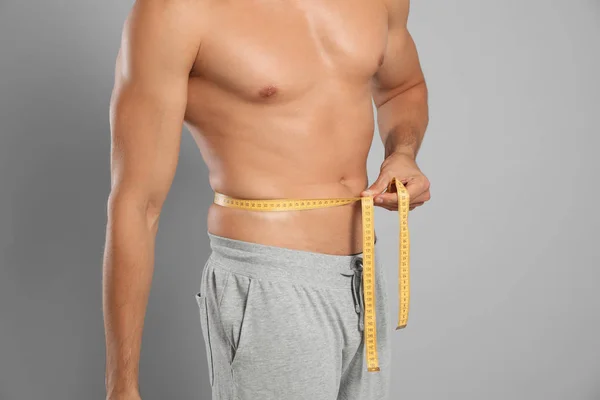 Young man with slim body using measuring tape on grey background, closeup view — Stock Photo, Image