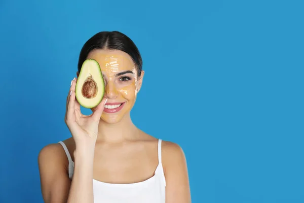 Young woman with cleansing mask on her face holding avocado against color background, space for text. Skin care