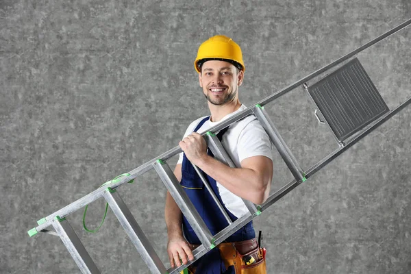 Handsome working man in hard hat holding ladder against color background