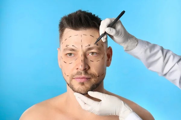 Doctor drawing marks on man\'s face for cosmetic surgery operation against blue background