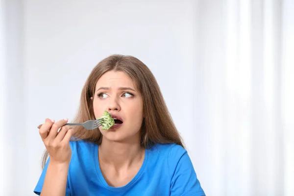 Portrait of unhappy woman eating broccoli on light background — Stock Photo, Image