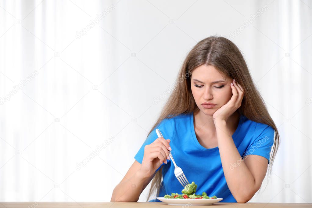 Portrait of unhappy woman eating broccoli salad at table on light background