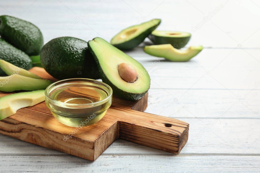 Board with bowl of natural oil and avocados on white wooden background. Space for text