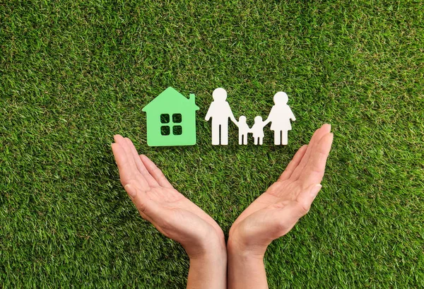 Mulher de mãos dadas perto de figuras de casa e família na grama verde, vista superior — Fotografia de Stock
