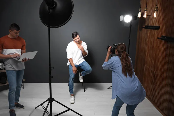 Fotógrafo profissional com assistente tirando foto de jovem no estúdio moderno — Fotografia de Stock