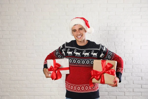 Happy man in Christmas sweater and Santa hat holding gift boxes near white brick wall — Stock Photo, Image