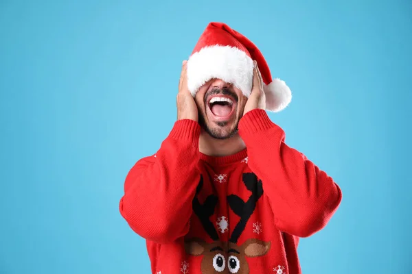 Emotional young man in Christmas sweater and Santa hat on light blue background — Stock Photo, Image