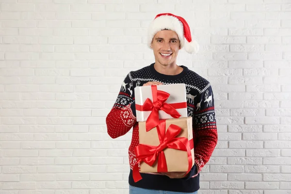Homem feliz em camisola de Natal e chapéu de Santa segurando caixas de presente perto da parede de tijolo branco — Fotografia de Stock