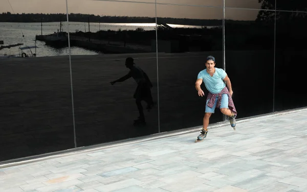 Handsome young man roller skating near glass building on city street, space for text