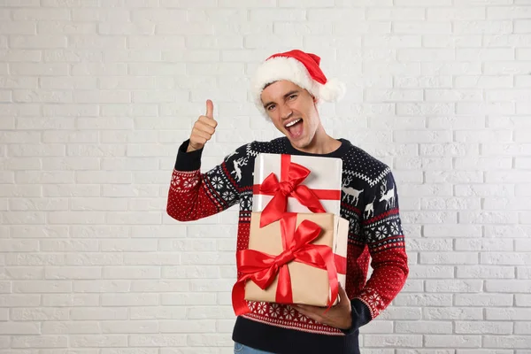 Happy man in Christmas sweater and Santa hat holding gift boxes near white brick wall — Stock Photo, Image