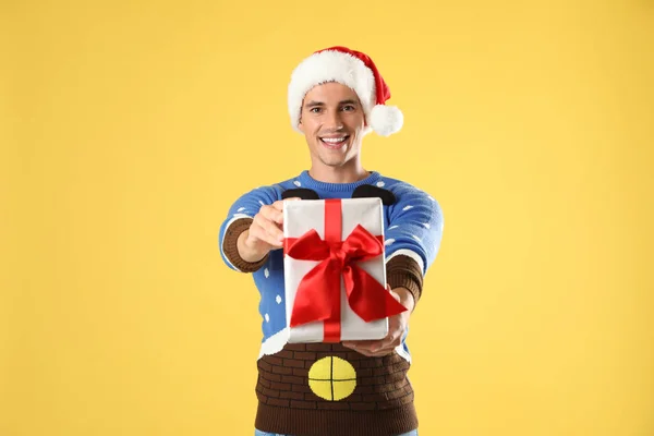 Happy man in Christmas sweater and Santa hat holding gift box on yellow background — Stock Photo, Image