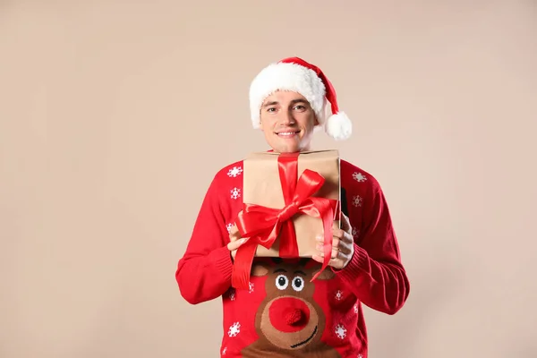 Happy man in Christmas sweater and Santa hat holding gift box on beige background — Stock Photo, Image