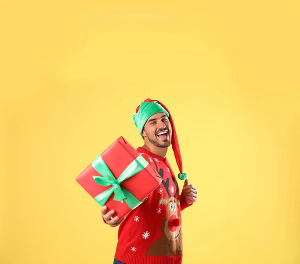 Happy man in Christmas sweater and elf hat holding gift box on yellow background — Stock Photo, Image