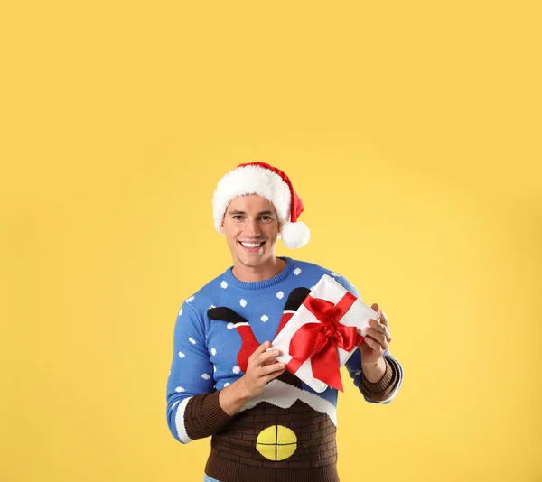 Happy man in Christmas sweater and Santa hat holding gift box on yellow background — Stock Photo, Image