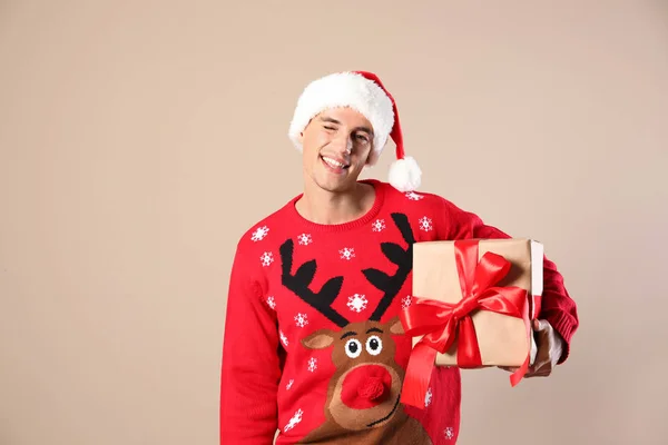 Homem feliz em camisola de Natal e chapéu de Santa segurando caixa de presente no fundo bege — Fotografia de Stock