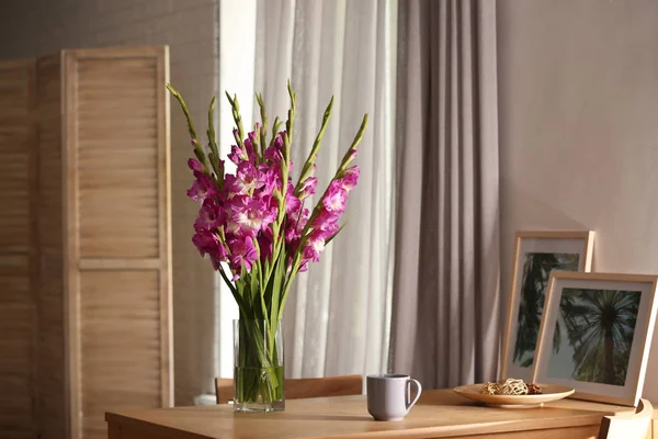 Vase with beautiful pink gladiolus flowers, pictures and cup on wooden table in room, space for text — Stock Photo, Image