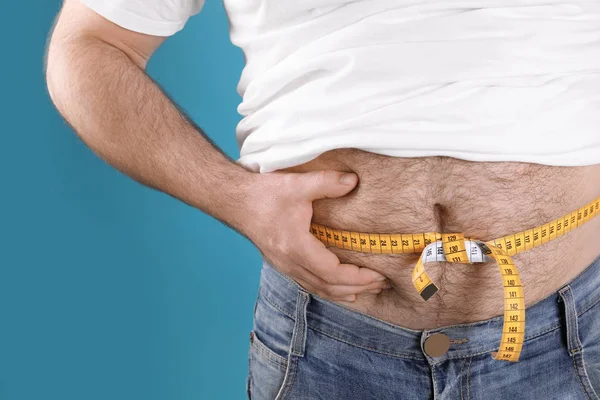 Overweight man with large belly and measuring tape on color background, closeup