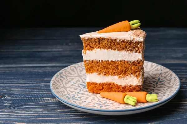 Piece of sweet carrot cake with delicious cream on blue wooden table against dark background. Space for text