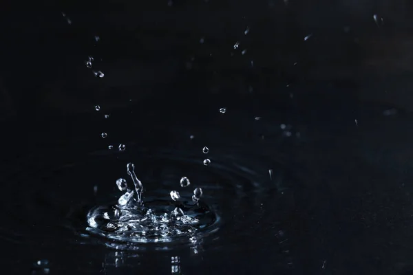 Rain drop falling down into puddle on dark background