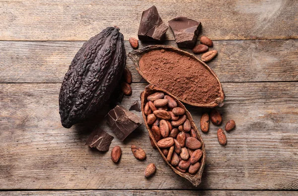 Cocoa pods with powder, beans and chocolate pieces on wooden table, flat lay