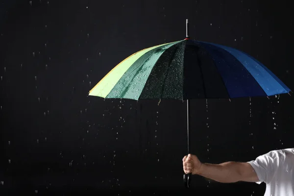 Hombre sosteniendo paraguas de color bajo la lluvia sobre fondo negro, primer plano — Foto de Stock