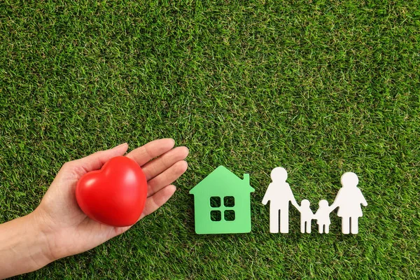 Mujer sosteniendo el corazón rojo en la mano cerca de figuras de la casa y la familia en la hierba verde, vista superior — Foto de Stock