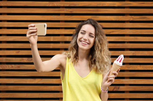 Glückliche junge Frau mit leckerem Eis in Waffelkegel macht Selfie in der Nähe der Holzwand — Stockfoto