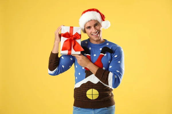 Happy man in Christmas sweater and Santa hat holding gift box on yellow background — Stock Photo, Image