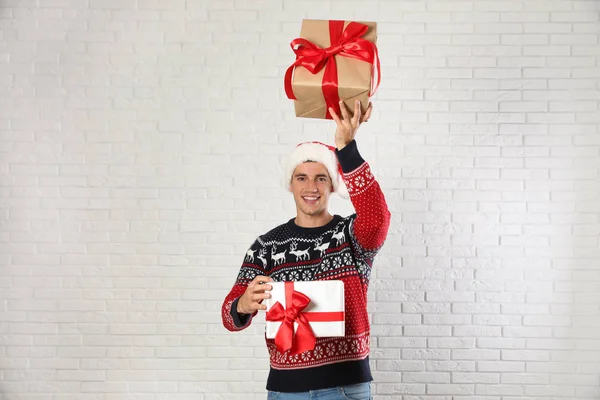 Homme heureux en pull de Noël et chapeau de Père Noël tenant des boîtes-cadeaux près du mur de briques blanches — Photo
