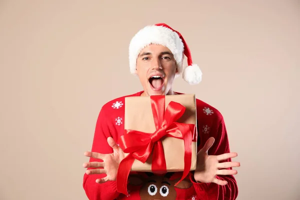Happy man in Christmas sweater and Santa hat holding gift box on beige background — Stock Photo, Image