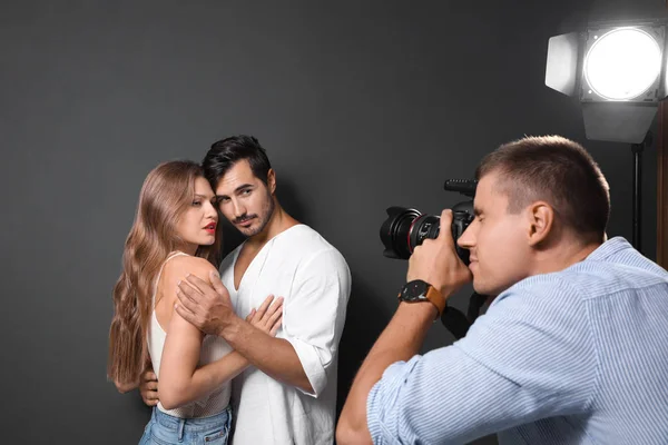 Fotógrafo profesional tomando fotos de parejas jóvenes sobre fondo gris oscuro en estudio moderno —  Fotos de Stock