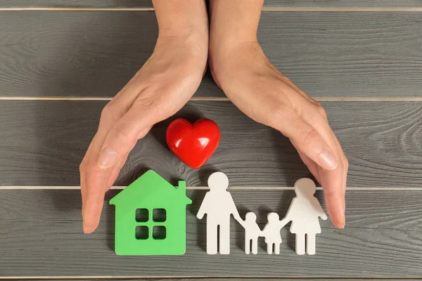 Vrouw Holding handen in de buurt van figuren van huis, rood hart en familie op grijze houten achtergrond, Top uitzicht — Stockfoto