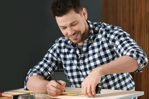 Handsome working man making marks on timber at table indoors. Home repair