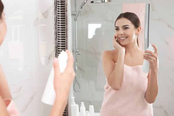 Ung kvinna som tillämpar termiskt vatten på ansiktet nära spegeln i Bad rums. Kosmetisk produkt — Stockfoto