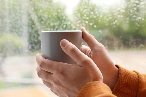 Junger Mann mit Tasse Kaffee am Fenster drinnen an einem regnerischen Tag, Nahaufnahme — Stockfoto