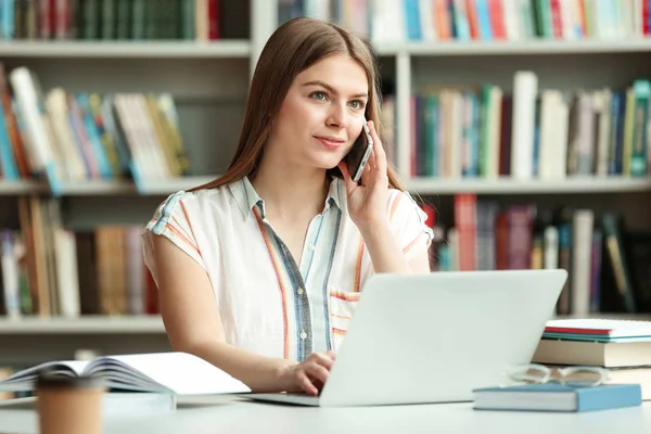 Jonge vrouw praten op telefoon en werken met laptop aan tafel in de bibliotheek — Stockfoto