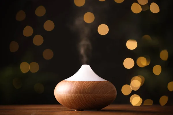 Modern essential oil diffuser on wooden table against blurred lights, space for text — Stock Photo, Image