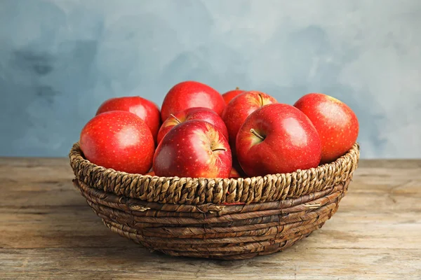 Wicker bowl with ripe juicy red apples on wooden table against blue background — Stock Photo, Image