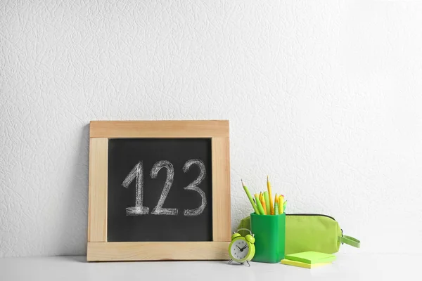 Kleine Tafel mit Zahlen und verschiedenen Schulschreibwaren auf Holztisch neben weißer Wand — Stockfoto