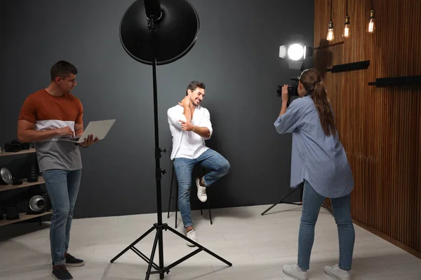 Fotógrafo profissional com assistente tirando foto de jovem no estúdio moderno — Fotografia de Stock